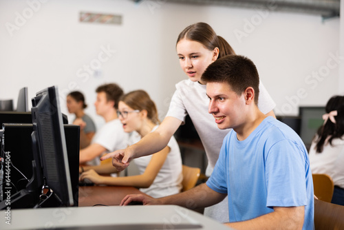 Friendly teen schoolgirl helping interested focused male classmate sitting at computer in information technology class of school