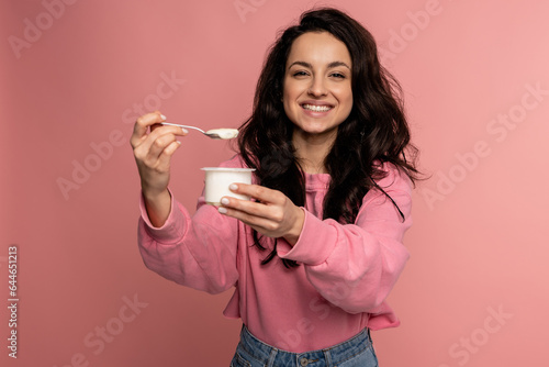 Waist-up portrait of a joyous brunette snacking on her favorite yogurt with the teaspoon from the plastic cup during the studio photo shoot photo