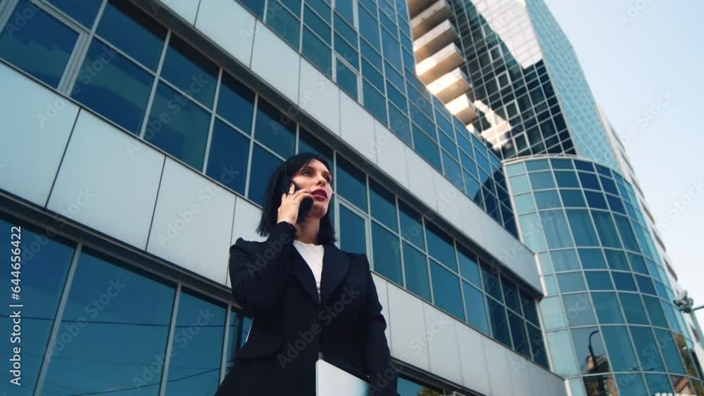 Successful elegant young business woman in a suit, standing in a big city and talking on a mobile phone, against the sky of city buildings, slow motion. Successful elegant young business woman in a suit, standing in a big city and talking on a mobile phone, against the sky of city buildings, slow motion.