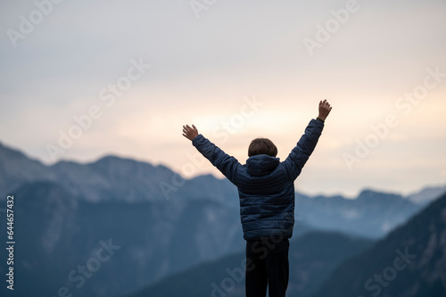 Toddler boy standing with his arms raised up high as he stands on top of the mountain looking