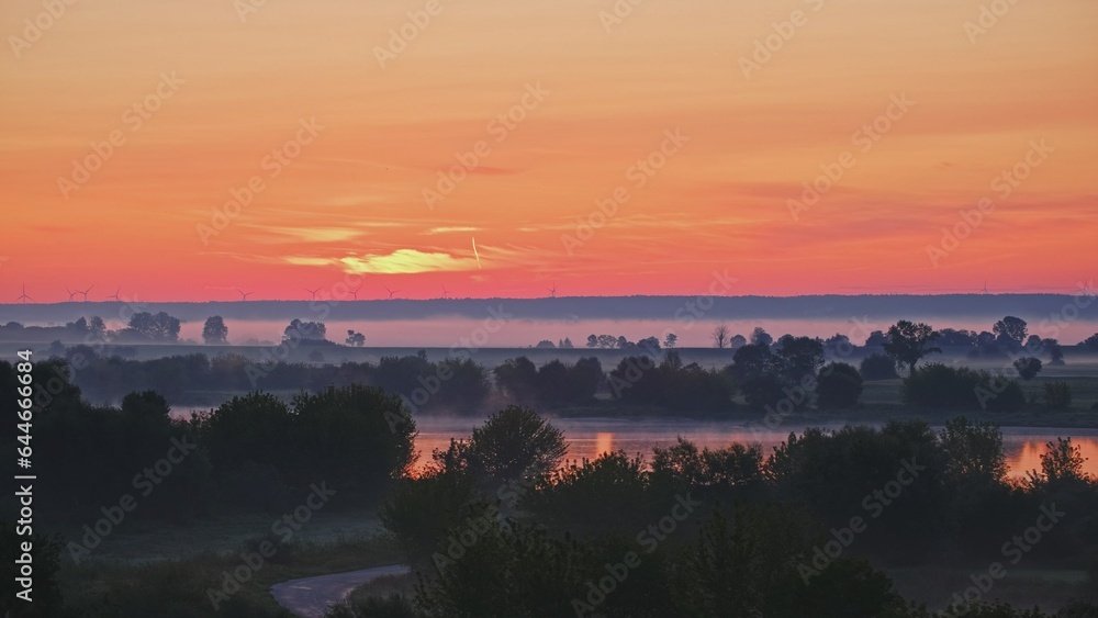 Beautiful Scenic Sunrise Morning Landscape with Fog Floating over River and Fields and Wind Turbines Rotating on Distatnt Hills on the Horizon 