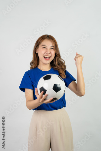 Happy Asian female soccer fan sending support to favorite team with soccer ball  smiling woman in blue t-shirt holding soccer ball to cheer at soccer game  isolated on white background.