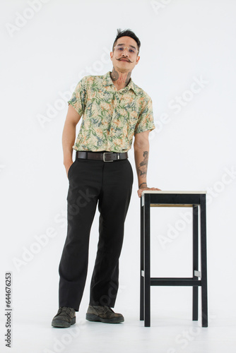 Isolated cutout studio full body shot Asian vintage classic male fashion model with mustache neck arm tattoo wearing eyeglasses in casual outfit standing near chair with boots on white background