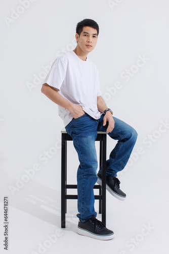 Portrait isolated cutout studio shot Asian handsome cool male fashion model in casual fashionable t shirt and blue jeans sitting on chair holding hand in pocket look at camera on white background