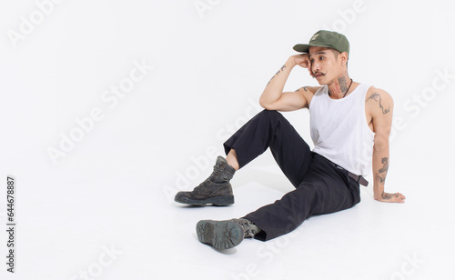 Isolated cutout studio shot Asian vintage classy mustache with neck arms hands tattoos male fashion model in casual fashionable sleeveless shirt cap boots sitting on floor on white background.