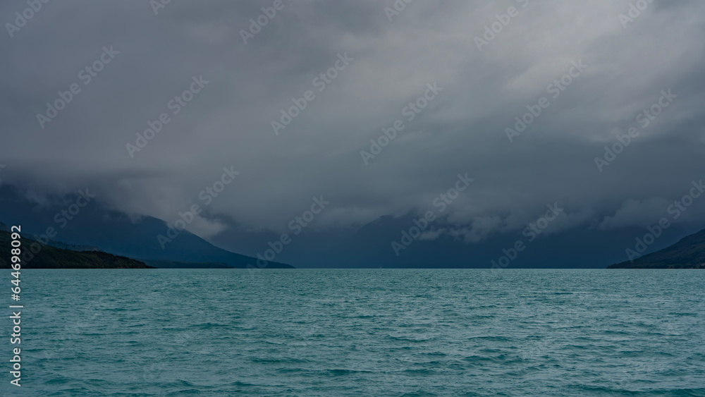 A beautiful turquoise glacial lake is surrounded by mountains. The peaks are hidden in fog and clouds. Ripples on the water. Argentina. Lago Argentino. El Calafate.