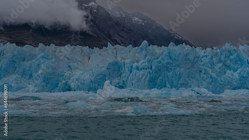 A wall of blue ice with sharp spikes rises above a turquoise lake. Broken icebergs and melting ice floes float in the water. Mountains in clouds and fog. Perito Moreno glacier. Argentina. El Calafate.