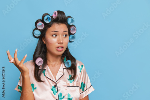 Portrait of a confused young asian woman in pajamas with curlers isolated over blue background