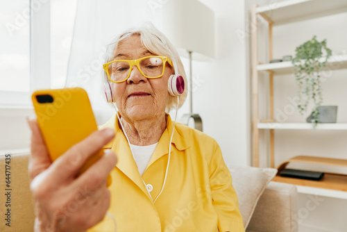 Happy elderly woman watching a video call on her phone and listening to music on her headphones, bright modern interior, lifestyle online communication.