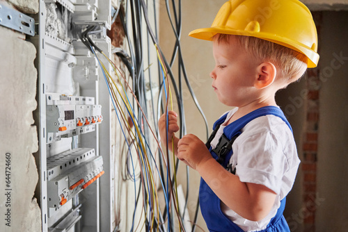 Little boy electrician playing with electrical wires while repairing electric switchboard or distribution board at home. Child checking electrical cables in apartment under renovation. photo
