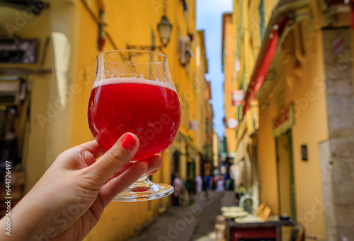 Woman s hand holding a glass of raspberry Lambic ale at an outdoor restaurant with a background of blurred buildings in old town Nice, South of France photo