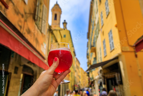 Fototapeta Naklejka Na Ścianę i Meble -  Woman s hand holding a glass of raspberry Lambic ale at an outdoor restaurant with a background of blurred buildings in old town Nice, South of France