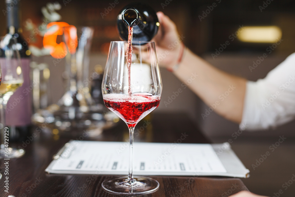 Faceless sommelier pours red wine into stem glass.