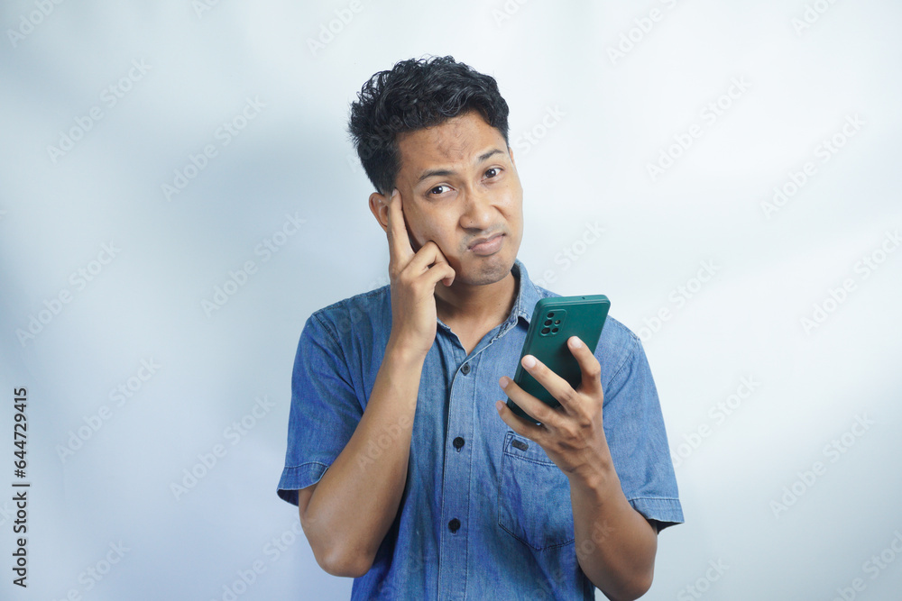 Adult Asian man looking to mobile phone that he hold with confused expression Adult Asian man looking to mobile phone that he hold with confused expression