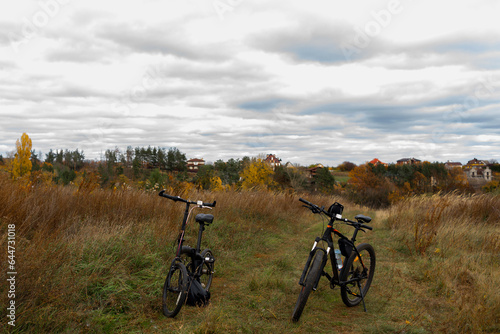 Two bicycles standing in autumn hill, meadow with brown grass. Fall country landscape with brown and orange forest trees, dried grass, sky with gray clouds. Travel cycling break, destination point.