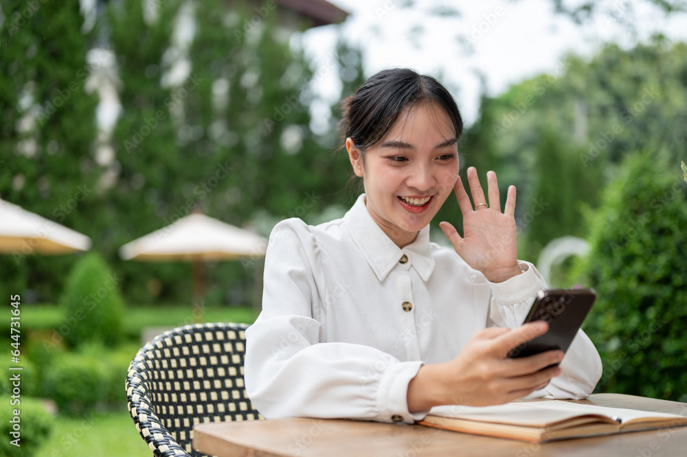 An attractive Asian woman enjoys talking with her friend on a video call while sitting in a garden. An attractive Asian woman enjoys talking with her friend on a video call while sitting in a garden.