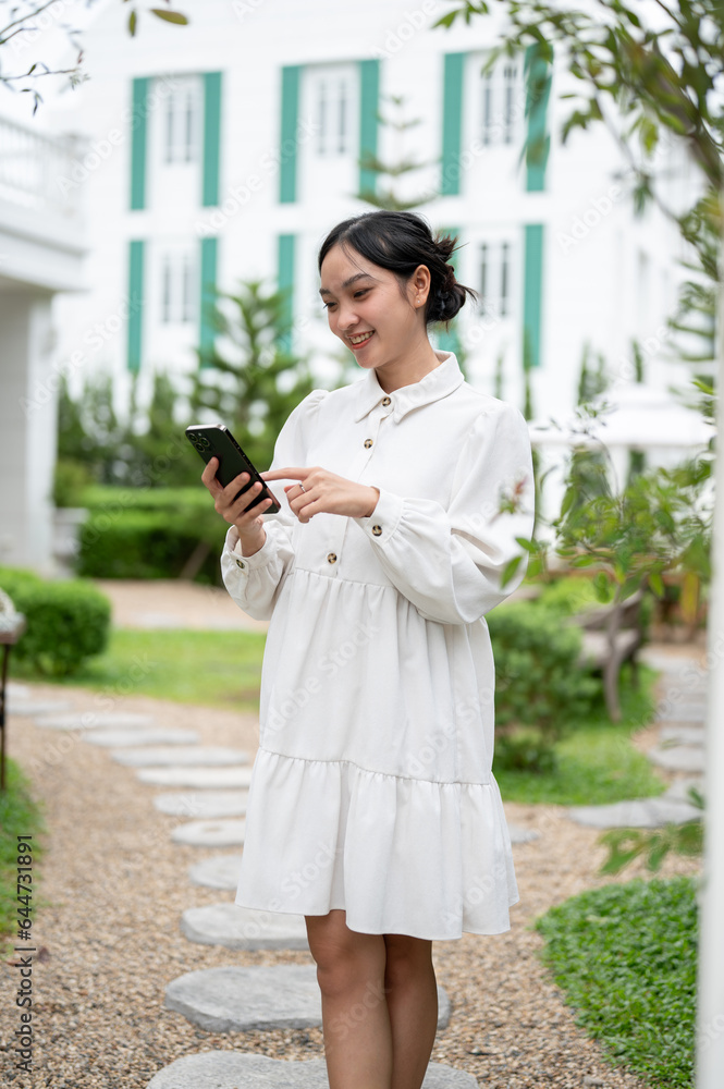 A happy Asian woman using her smartphone while strolling in her beautiful backyard garden. A happy Asian woman using her smartphone while strolling in her beautiful backyard garden.