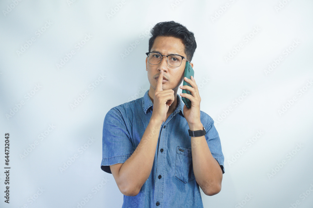 Asian man wearing blue shirt hides a secret while talking on the phone with finger on lips against white background. Asian man wearing blue shirt hides a secret while talking on the phone with finger on lips against white background.