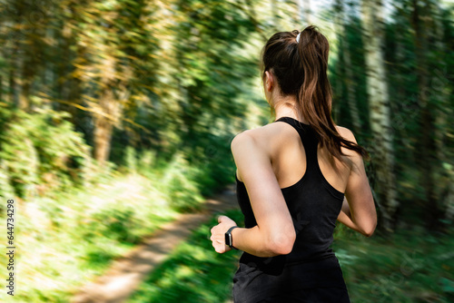 Young fitness woman in black sportwears running at morning forest trail. Rear view.