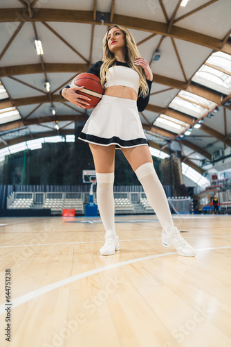 Low angle shot of young woman in a black and white cheerleader uniform standing on court indoor with a basketball . High quality photo