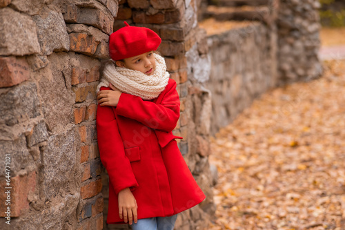 A sad Caucasian girl in a red coat and beret stands leaning against a brick wall. 
