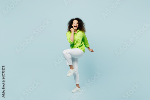 Full body young latin woman she wears green shirt casual clothes doing winner gesture celebrate clenching fists say yes isolated on plain pastel light blue cyan background studio. Lifestyle concept.