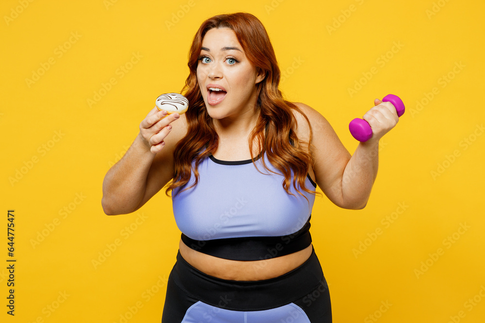 Young chubby overweight plus size big fat fit woman wearing blue top warm up training hold dumbbells eat donut look camera isolated on plain yellow background studio home gym. Workout sport concept. Young chubby overweight plus size big fat fit woman wearing blue top warm up training hold dumbbells eat donut look camera isolated on plain yellow background studio home gym. Workout sport concept.