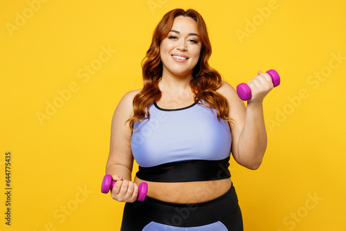 Young smiling happy chubby overweight plus size big fat fit woman wear blue top warm up training hold dumbbells look camera isolated on plain yellow background studio home gym. Workout sport concept.