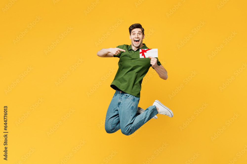Full body young happy man he wearing green t-shirt casual clothes jump high hold point finger on gift certificate coupon voucher card for store isolated on plain yellow background. Lifestyle concept.