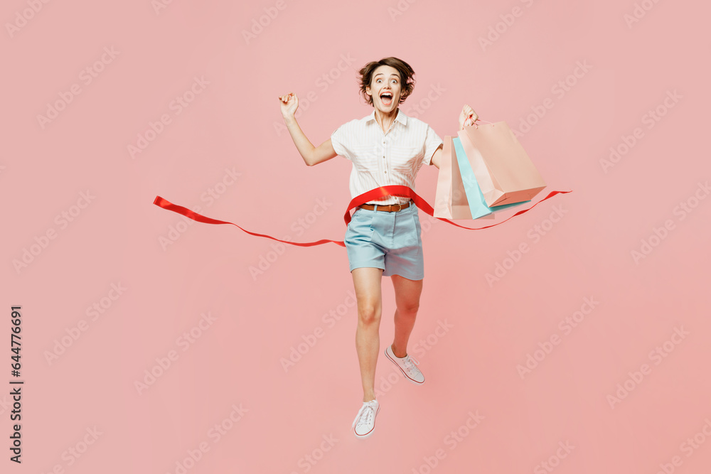 Full body overjoyed fun young woman wear casual clothes hold shopping paper package bags run cross finish red ribbon isolated on plain pink color background studio. Black Friday sale buy day concept. Full body overjoyed fun young woman wear casual clothes hold shopping paper package bags run cross finish red ribbon isolated on plain pink color background studio. Black Friday sale buy day concept.