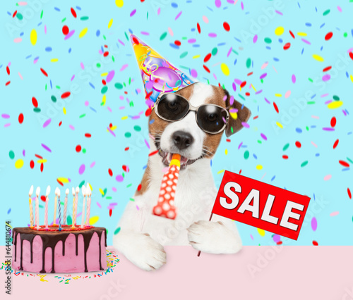 jack russell terrier puppy wearing sunglasses and party cap blows into party horn and showing signboard with labeled "sale" © Ermolaev Alexandr