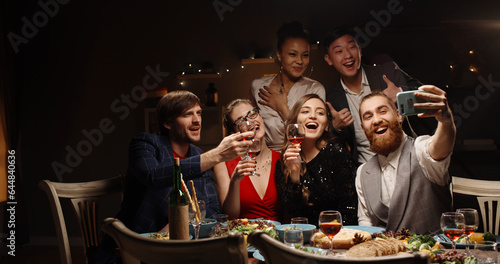 Group of cheerful multiethnic students celebrating a holiday together at dinner party  taking a group selfie and positively smiling - celebration  communication concept