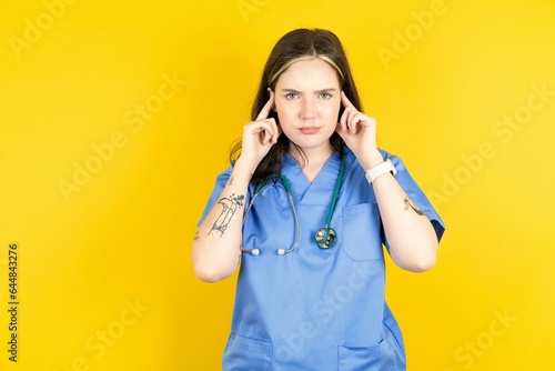 Serious concentrated Young caucasian doctor woman wearing blue medical uniform keeps fingers on temples, tries to ease tension, gather with thoughts and remember important information for exam photo