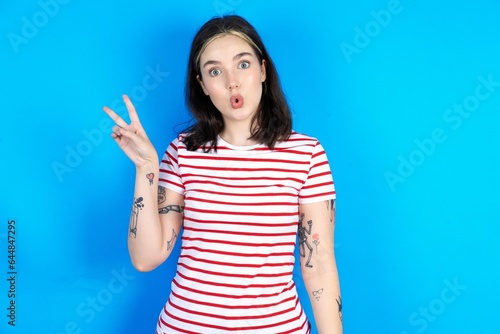 beautiful young woman wearing striped T-shirt makes peace gesture keeps lips folded shows v sign. Body language concept photo