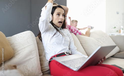 Woman in headphones and with laptops in her hands sits on sofa from behind  children play. Remote work in coronavirus in family concept