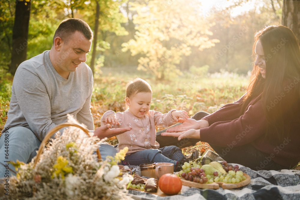 Fototapeta premium Mother, father, kid have picnic in autumn forest in nature. Mom, dad hugs daughter child sitting on blanket on yellow leaves in park. Family spending time together at sunset on vacation. Happy holiday