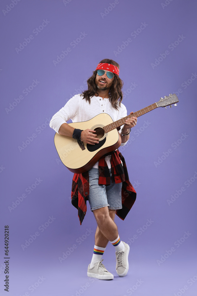 Stylish hippie man in sunglasses playing guitar on violet background