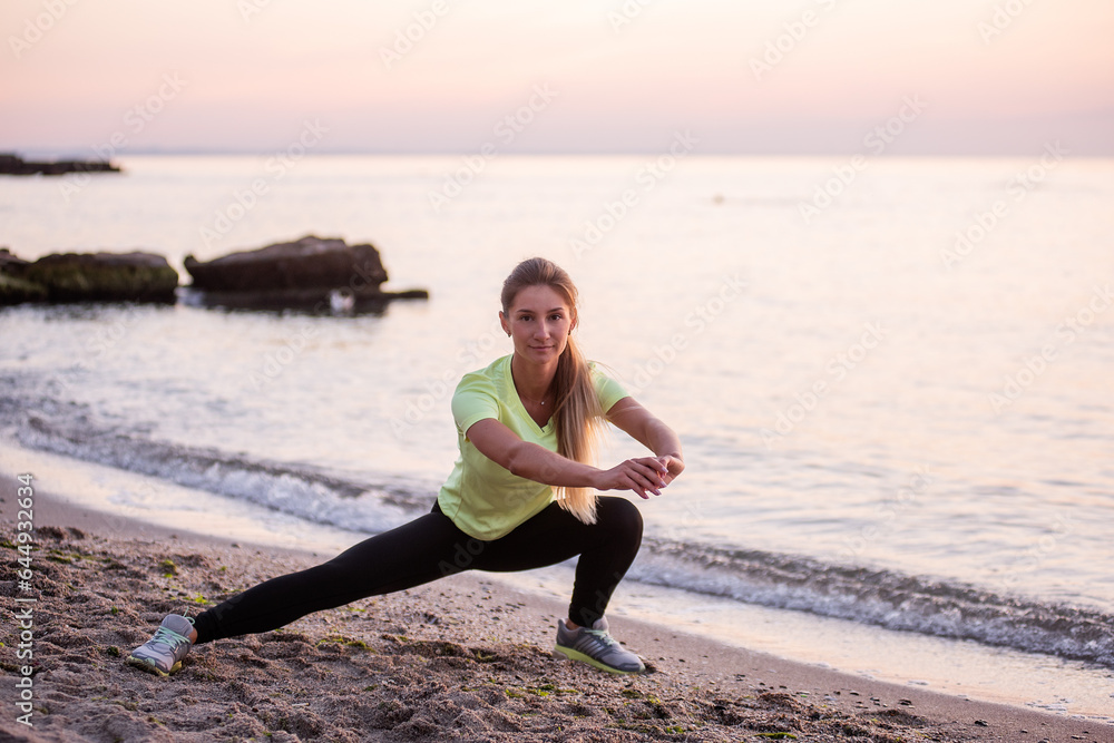 Young woman in sportswear does sit-ups early in morning on seashore at sunrise. Girl of slim build with long hair is stretching. Outdoor sports by the water. Fitness classes, body care, mental health Young woman in sportswear does sit-ups early in morning on seashore at sunrise. Girl of slim build with long hair is stretching. Outdoor sports by the water. Fitness classes, body care, mental health