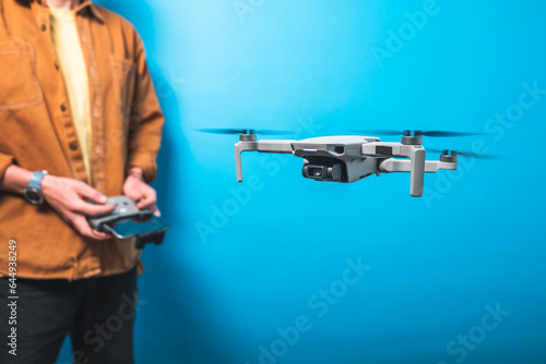 Smiling young man piloting a drone in flight training with a remote control on an isolated blue background. Drone pilot and aerial filming concepts. Focus on the drone