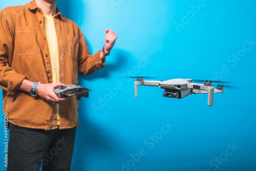 Smiling young man celebrating piloting a drone in flight with a remote control on an isolated blue background. Drone pilot and aerial filming concepts.