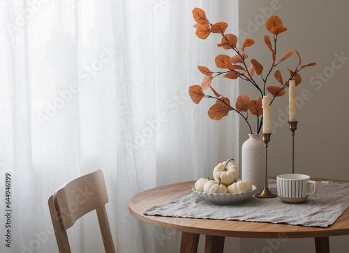 Autumn home interior in the living room - around wooden table, a chair, a bouquet of autumn leaves in a vase, decorative pumpkins. Cozy home