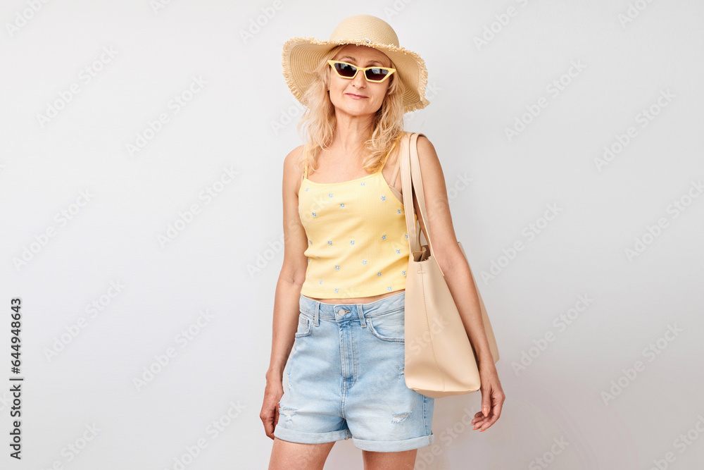 Portrait of happy beautiful blonde mature woman in yellow summer clothes, straw hat, sunglasses and shopping bag smiling isolated on white studio background. Vacation and travel concept Portrait of happy beautiful blonde mature woman in yellow summer clothes, straw hat, sunglasses and shopping bag smiling isolated on white studio background. Vacation and travel concept