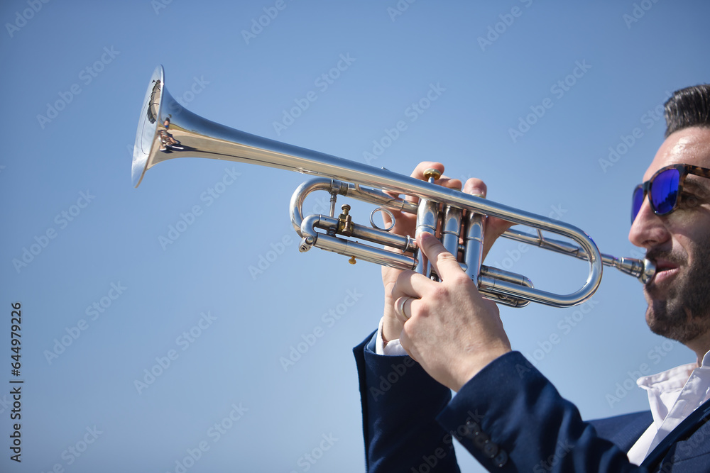 Obraz premium Young Hispanic man, wearing a jacket and sunglasses, playing a pretty, silvery trumpet outdoors. Concept, music, instruments, trumpet.