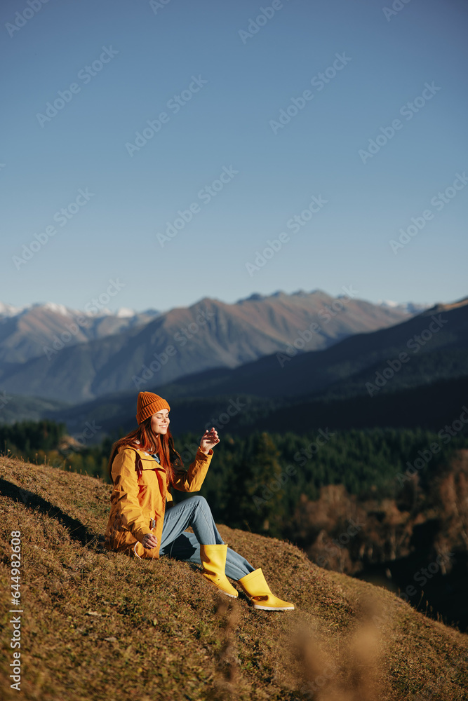 Fototapeta premium Woman full-length sitting resting on a hill and looking at the mountains in a yellow raincoat and jeans happy camping trip in the autumn, freedom lifestyle 
