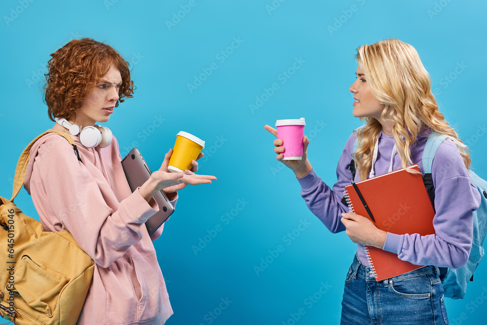 displeased teen friends with backpacks and paper cups pointing at each other and quarreling on blue displeased teen friends with backpacks and paper cups pointing at each other and quarreling on blue