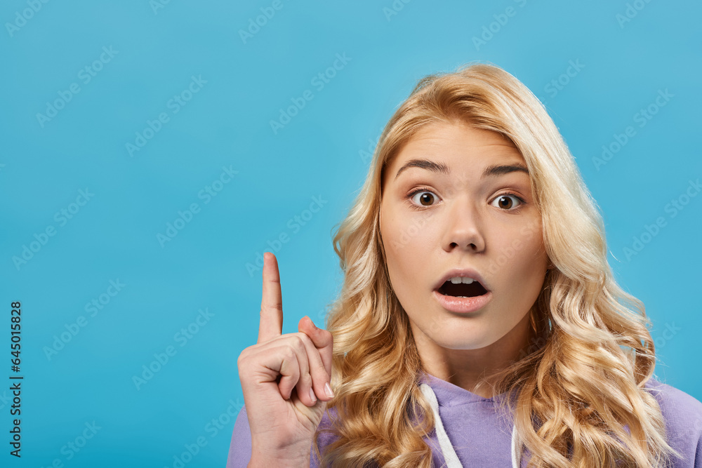 portrait of amazed blonde teenage girl with open mouth showing idea gesture on blue
