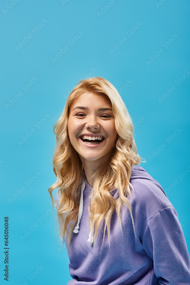 excited blonde teenage girl in hoodie looking at camera and laughing on blue, happiness excited blonde teenage girl in hoodie looking at camera and laughing on blue, happiness