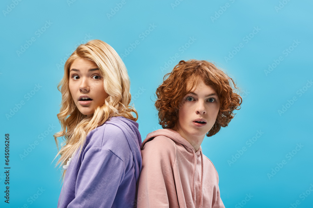 amazed redhead teen boy with blonde girlfriend sitting back to back and looking at camera on blue amazed redhead teen boy with blonde girlfriend sitting back to back and looking at camera on blue