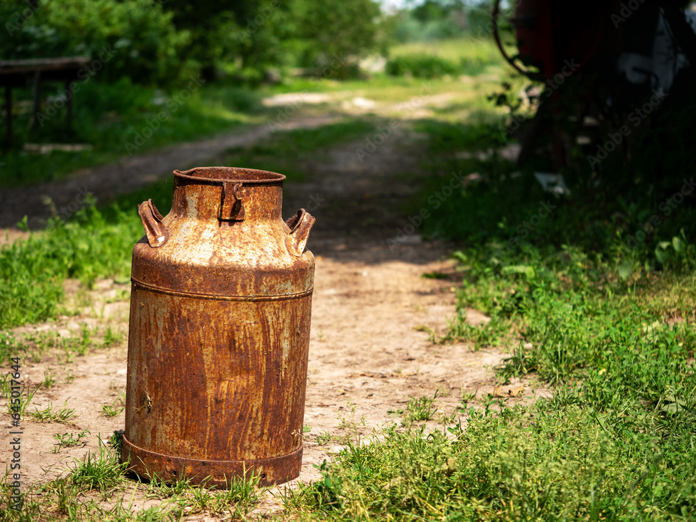 Old rusty metal milk churn standing on a footpath or country road. Vintage antique container for liquid. Rural country side life concept. Old rusty metal milk churn standing on a footpath or country road. Vintage antique container for liquid. Rural country side life concept.