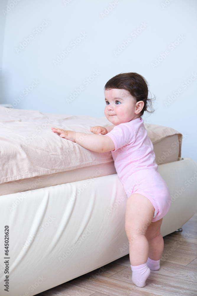 Cute baby girl in bodysuit stand holding bed frame, learning to walk Cute baby girl in bodysuit stand holding bed frame, learning to walk